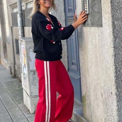 Mulher de casaco preto e calças vermelhas com riscas brancas numa rua urbana junto a porta cinzenta