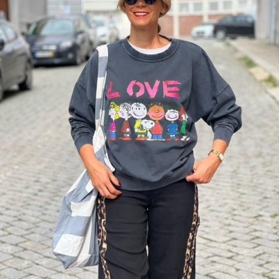 Mulher vestindo sweatshirt preta com estampa dos Peanuts e palavra LOVE, boné preto e calças pretas com faixa lateral de leopardo num ambiente urbano.