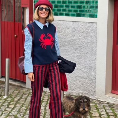 Mulher com roupa colorida e cão ao lado em rua de pedra com azulejos verdes e porta vermelha.