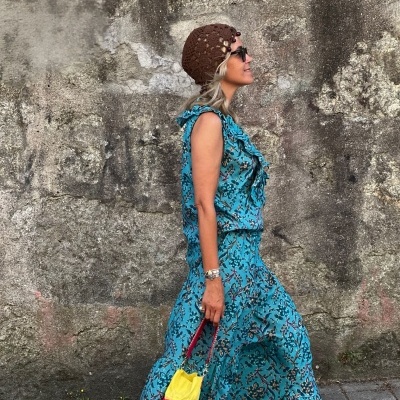 Mulher com vestido azul longo floral e bolsa colorida junto a parede de pedra