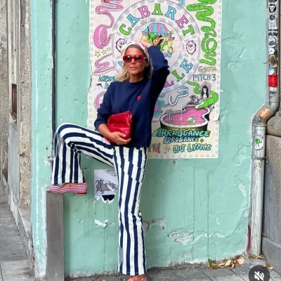 Mulher com roupa azul e branca e óculos de sol vermelhos junto a parede verde com cartaz colorido
