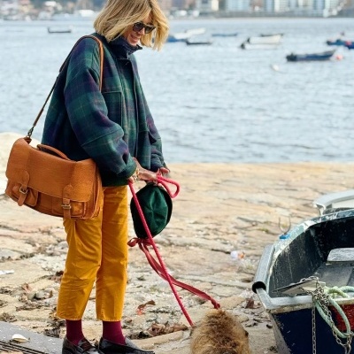 Mulher com saco de pele castanha e roupa colorida ao lado de cão perto de um barco