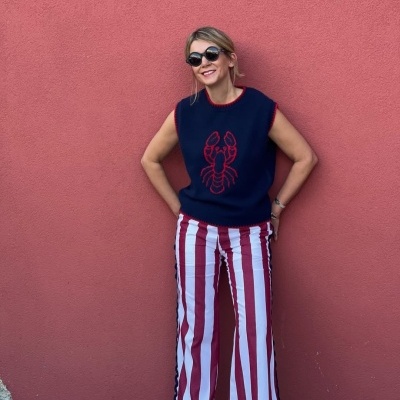 Mulher com suéter azul marinho e calças às riscas junto a parede rosa