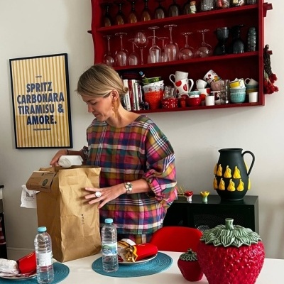 Mulher com blusa xadrez desembrulha saco de papel numa cozinha com prateleira vermelha cheia de copos e canecas, e duas garrafas de água na mesa