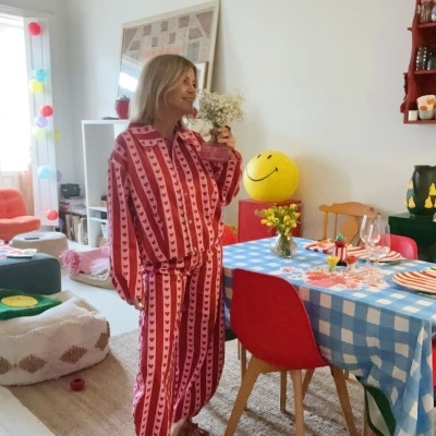 Mulher com pijama rosa e vermelho com corações, segurando flores brancas, em sala de jantar colorida.