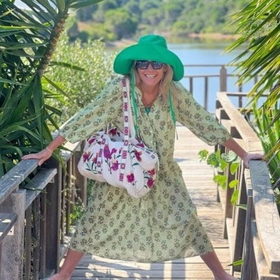 Mulher com vestido verde claro florido, chapéu verde e mala branca floral numa ponte de madeira junto a um lago