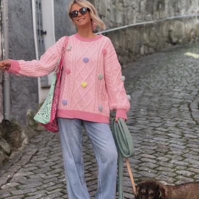 Mulher com suéter rosa de malha e pompons, calças largas azuis, ténis e óculos escuros a passear cão castanho em rua de pedra