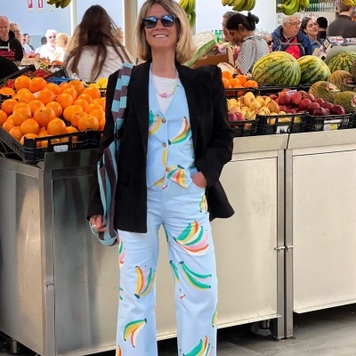 Mulher vestida com conjunto azul claro com padrão de bananas coloridas e casaco preto num mercado de frutas