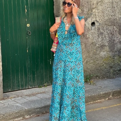 Mulher com vestido longo azul floral, acessório de cabeça castanho, óculos e bolsa colorida em rua com porta verde e parede cinzenta