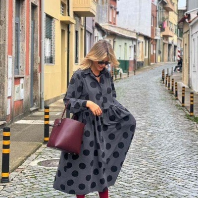 Mulher com vestido comprido cinzento de bolinhas pretas e mala bordeaux numa rua pavimentada.