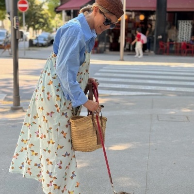 Mulher vestindo vestido estampado com cães, camisa azul, chapéu castanho, segurando bolsa de palha e trela vermelha com cão peludo em passeio na rua