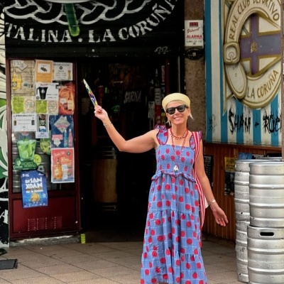 Mulher com vestido azul e bolas vermelhas, chapéu bege e sapatos vermelhos, em frente a estabelecimento com cartazes e barris.