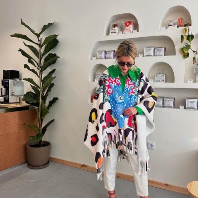 Pessoa com manta colorida abstrata, calças brancas e sapatos vermelhos em interior com prateleiras e planta