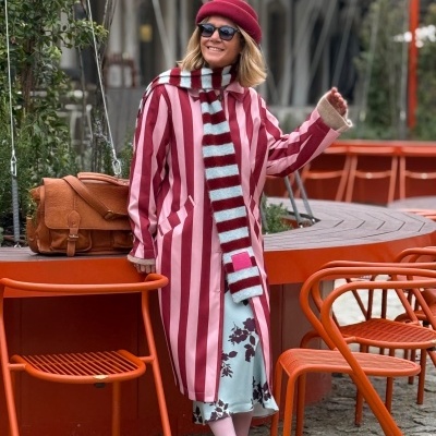 Mulher com casaco às riscas rosa e vermelho, cachecol listrado, boné vermelho e saia azul clara com flores em cadeiraria urbana