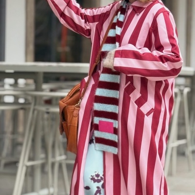 Mulher com casaco comprido às riscas rosa e vermelho, cachecol azul e vermelho, mala castanha e vestido azul com padrão roxo.