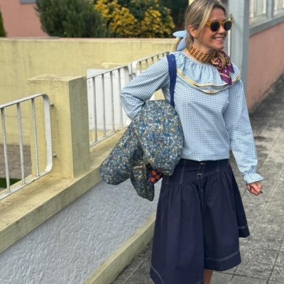 Mulher vestida com blusa azul clara xadrez e saia azul escura segurando casaco floral e mochila azul numa calçada