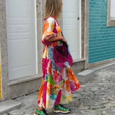 Mulher com vestido floral colorido, sapatos verdes e bolsa vinho a caminhar em rua de calçada de pedras