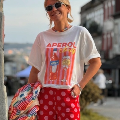 Mulher com t-shirt branca estampada, calças vermelhas com bolinhas, acessórios coloridos e óculos escuros em ambiente urbano