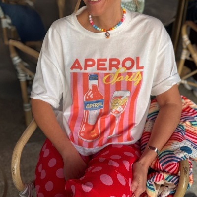 Pessoa com t-shirt branca estampada APEROL Spritz, calças vermelhas com bolinhas rosa e colar colorido sentada numa cadeira de vime