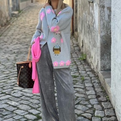 Mulher com casaco de malha cinzento com flores cor-de-rosa e cupcake, calças largas cinzentas, sapatos amarelos, lenço cor-de-rosa, chapéu cinzento e óculos de sol numa rua de pedra