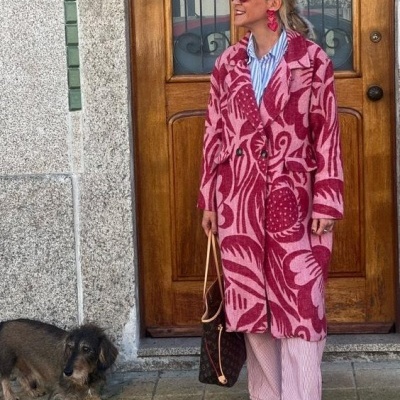 Mulher com casaco rosa e vermelho com padrão floral e calças cor-de-rosa em frente a porta de madeira com cão pequeno castanho ao lado.