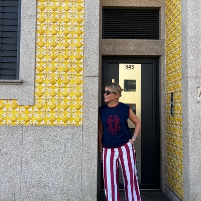 Mulher com camisa azul e calças riscas vermelhas junto a porta preta com número 343 e azulejos amarelos florais.