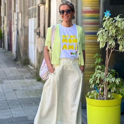 Mulher com t-shirt branca com texto amarelo, camisola amarela, saia creme, sapatilhas cinzentas, óculos de sol, parada junto a vaso de planta amarelo