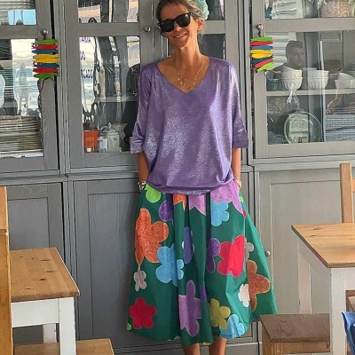 Mulher vestindo saia florida verde e camisola lilás em ambiente de cozinha com móveis em madeira e armários cinza