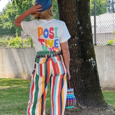 Pessoa de costas com roupa colorida e chapéu azul junto a uma árvore ao ar livre.