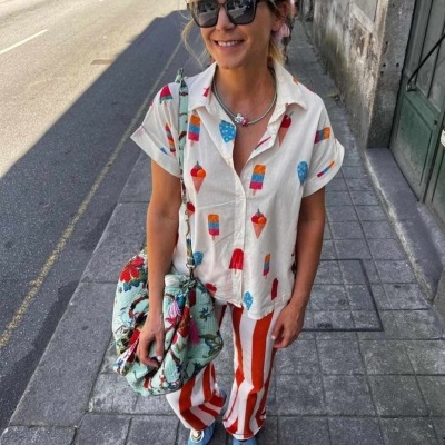 Mulher na rua com camisa estampada de picolés, calças às riscas vermelhas e brancas, sapatos azuis e óculos escuros