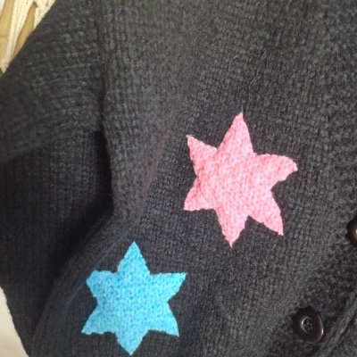 Casaco de malha preta com estrelas de feltro azul e rosa e parte amarela, pendurado num cabide