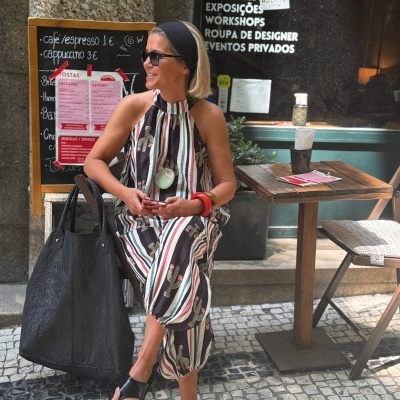 Mulher com vestido às riscas e padrão de cactos, sandálias pretas e acessórios vermelhos sentada em esplanada ao ar livre