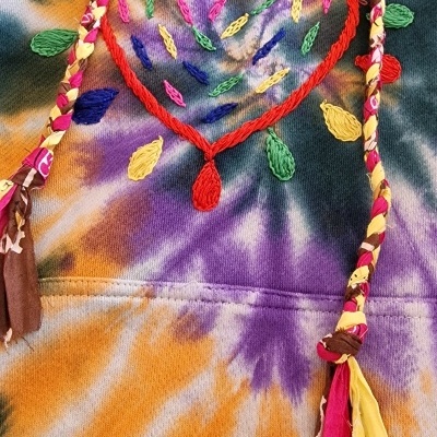 Detalhe de roupa tie-dye com bordados coloridos e cordões trançados