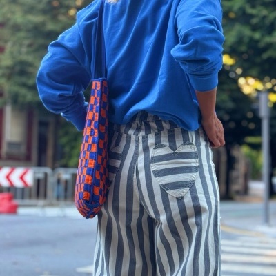 Pessoa de costas com camisola azul e calças riscas brancas e cinzentas, bolsa xadrez laranja e azul no ombro, em rua urbana.
