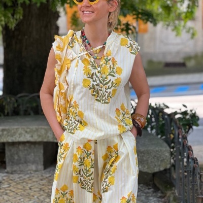 Mulher com conjunto floral amarelo e branco, óculos de sol amarelos e acessórios, ao ar livre junto a árvore e banco de pedra.