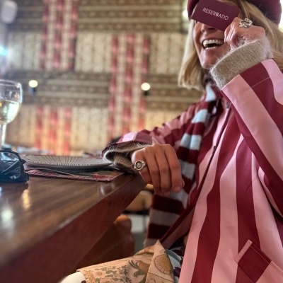 Mulher com blazer riscado rosa e vermelho, boina e cachecol segurando cartão RESERVADO ao lado de mesa de madeira.