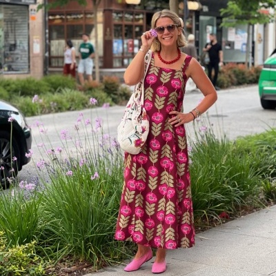 Mulher com vestido comprido floral vermelho e rosa, sapatos rosa, colar vermelho e óculos de sol, em rua com flores e árvores