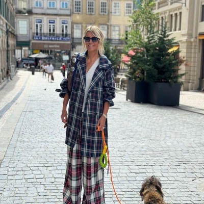 Mulher com casaco e calças xadrez a passear cão pequeno de pelo castanho numa rua de calçada em cityscape