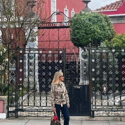 Mulher com blusa animal print e mala marrom à frente de portão preto decorativo e prédio vermelho.