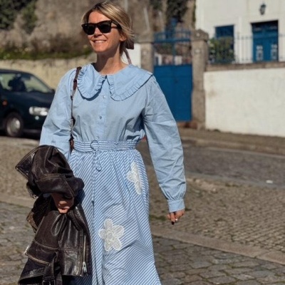 Mulher de vestido azul claro com padrão riscado e flores brancas, segurando jaqueta preta na mão