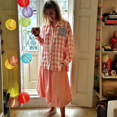 Mulher com camisa xadrez rosa e saia rosa segurando caneca branca com círculos vermelhos perto de janela com cortinas de papel coloridas