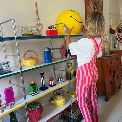 Estante com objetos decorativos coloridos e mulher com calças de riscas a segurar bola sorridente amarela
