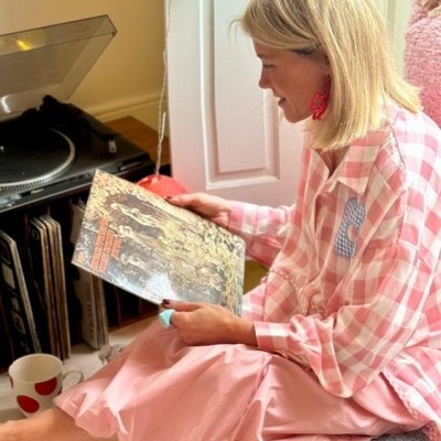 Pessoa sentada com camisa xadrez e saia rosa a segurar disco de vinil junto a gira-discos
