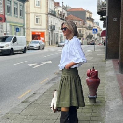 Mulher com blusa branca, saia verde, botas pretas e óculos escuros numa rua urbana