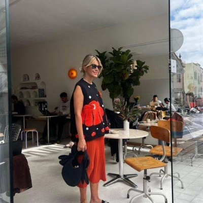 Mulher num colete preto com padrão vermelho, saia vermelha e sapatos pretos numa cafetaria moderna