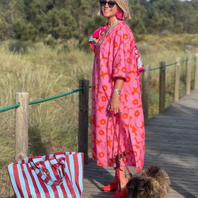 Mulher com vestido longo floral rosa e laranja e acessórios, com saco listrado e cão num passadiço
