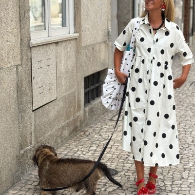 Mulher com vestido branco estampado a preto e sandálias vermelhas a passear com cão numa calçada portuguesa