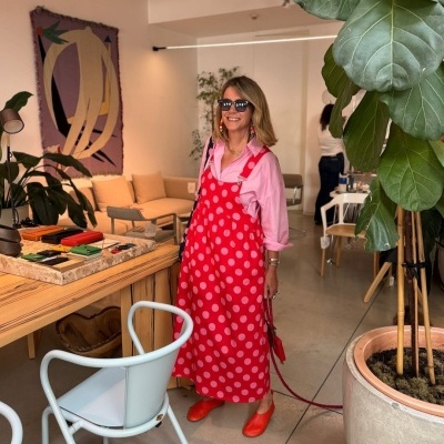 Mulher vestindo vestido vermelho de bolinhas rosa e camisa rosa, sapatos laranja e óculos escuros, em ambiente interior decorado com plantas e móveis de madeira