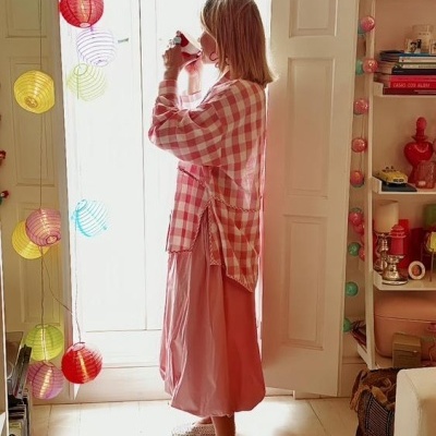 Mulher com blusa xadrez vermelho e branco e saia rosa junto a porta branca com lanternas de papel coloridas