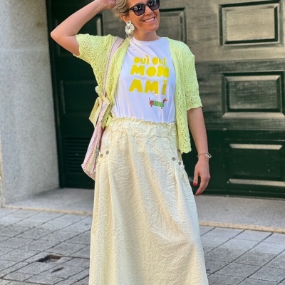 Mulher com vestido amarelo claro, camiseta branca com texto amarelo, cardigan amarelo, brincos grandes, óculos escuros e bolsa bege.
